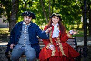 Two people in 18th-century-style costumes sit on a bench outdoors. Featured in this Estonia Travel Blog, the man wears a blue coat with silver buttons, and the woman wears a red coat with gold trim; both sport tricorne hats and share a slight smile.