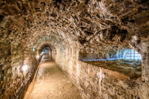 A long, arched stone tunnel in the Casemates of Victoria Bastion in Narva, with rough, uneven walls and ceiling, is illuminated by lights on the left. The sandy floor is worn, and an iron-barred alcove on the right lets light stream in.