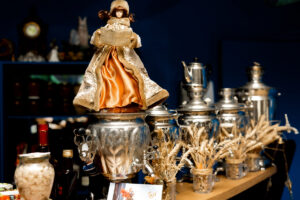 A vintage-style doll in a gold and orange dress sits atop a polished metal samovar, evoking the charm of a Culinary Cultural Tour to Eastern Estonia. Surrounding it are samovars, dried wheat, bottles, and preserves on a wooden shelf against a dark blue backdrop.