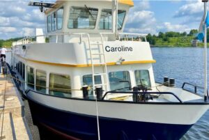 A white and blue boat named “Caroline” is docked at a pier on a sunny day, ready for its Mining Museum Tour. The boat has two decks with large windows, a yellow stripe, and a flag at the stern, set against green trees and a blue sky with clouds.