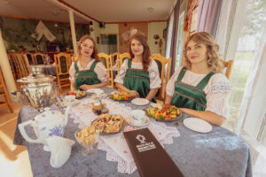 Three women in traditional green and white dresses sit around a table set for Russian tea, delicious pastries, and local dishes—a scene from a Culinary Cultural Tour to Eastern Estonia—complete with a samovar and menu in a cozy, well-lit room. Kolkja fish and onion restaurant, traditional Peipsi dishes