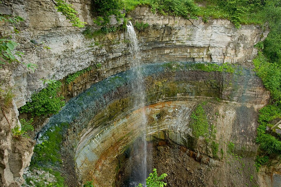 Tall, thin Valaste waterfall spills from a rocky cliff into a circular gorge. The layered rock walls display shades of gray, green, and brown, with lush vegetation clinging to ledges and rim, as mist rises from the falls to the rocky floor below.
